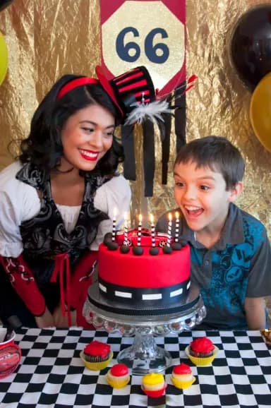 A Happily Ever Laughter performer and a delighted child admire a candle-lit race car birthday cake at a themed birthday party.