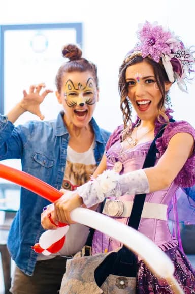Two adults laugh together, one with playful cat face paint, while a Happily Ever Laughter performer in a purple costume twists balloon animals at a company event.