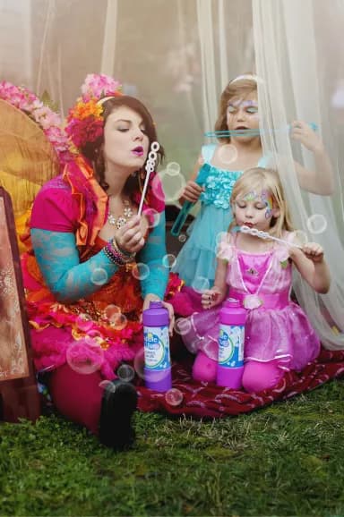 A Happily Ever Laughter faerie blows bubbles with two young children in fairy costumes at an elegant outdoor garden party.