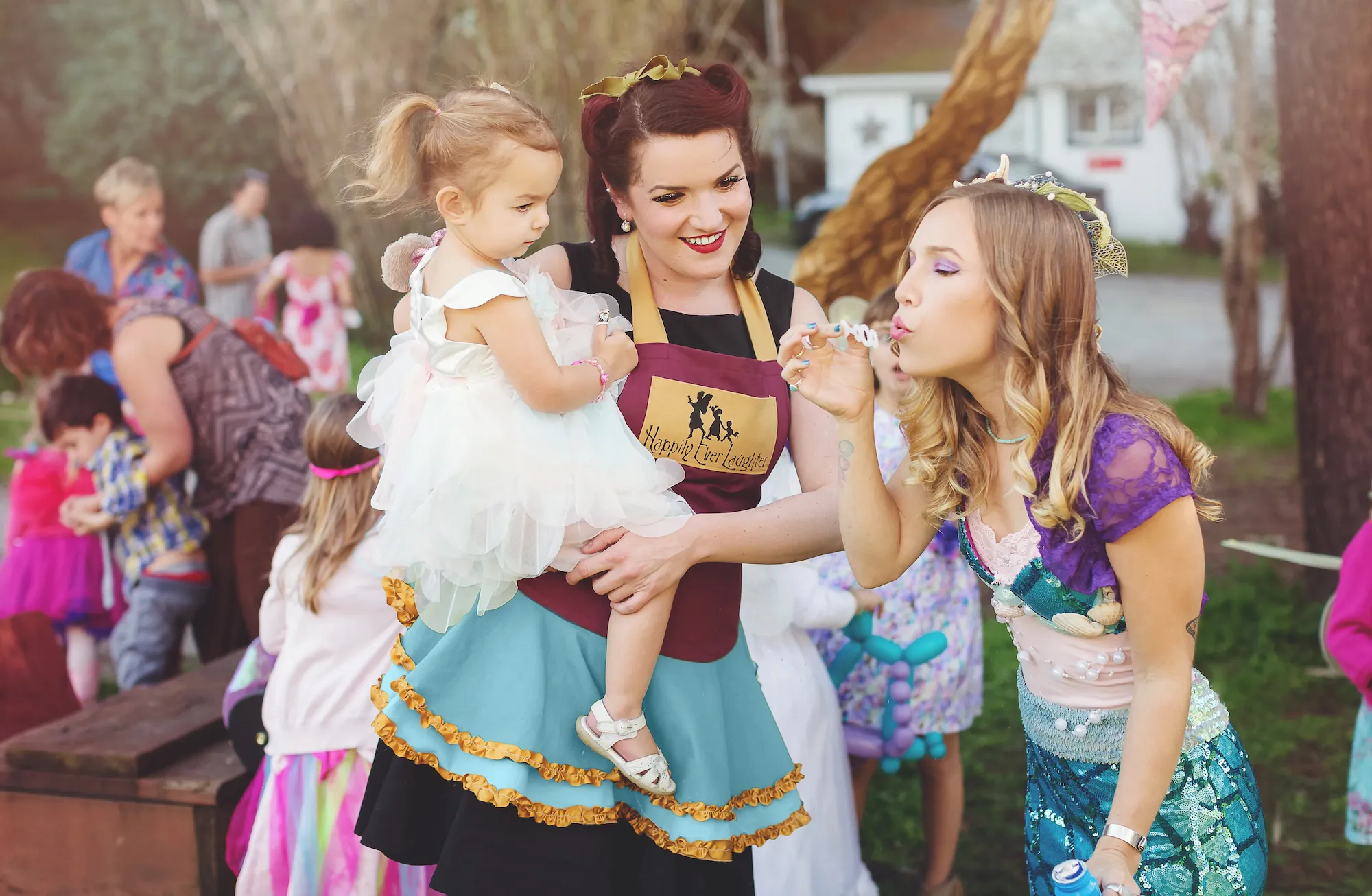 A Happily Ever Laughter party faerie holds a small child in a fairy costume while a performer in a mermaid costume plays with her at an outdoor children's party.