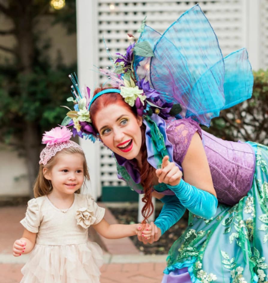 A Happily Ever Laughter faerie kneeling beside a young child in a flower-crown costume at an outdoor party.