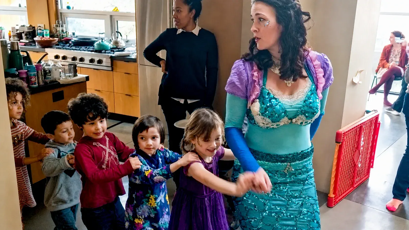 A Happily Ever Laughter faerie performer in a teal gown leading a long line of children in a follow-the-leader parade through a home.