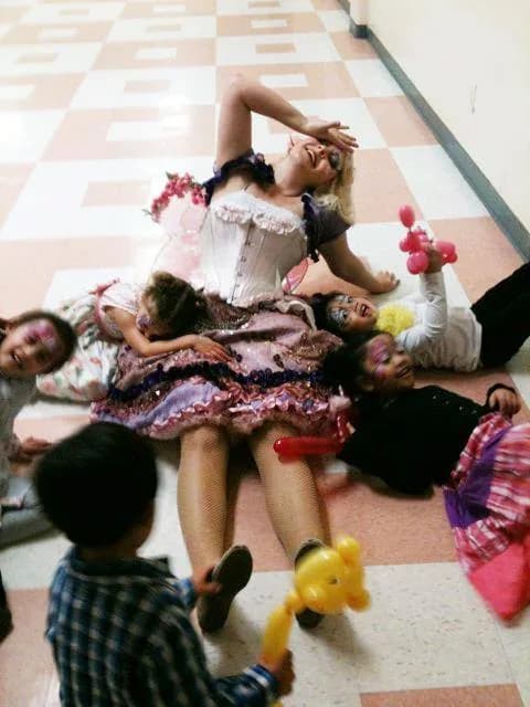 A Happily Ever Laughter faerie performer lies on a tile floor surrounded by children holding balloon animals at the end of a party.