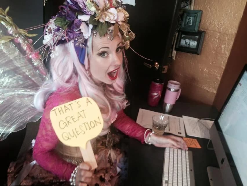 Fae Gershenson, founder of Happily Ever Laughter Parties, in a pink dress and floral faerie crown at her desk, holding a sign that reads That's a great question.