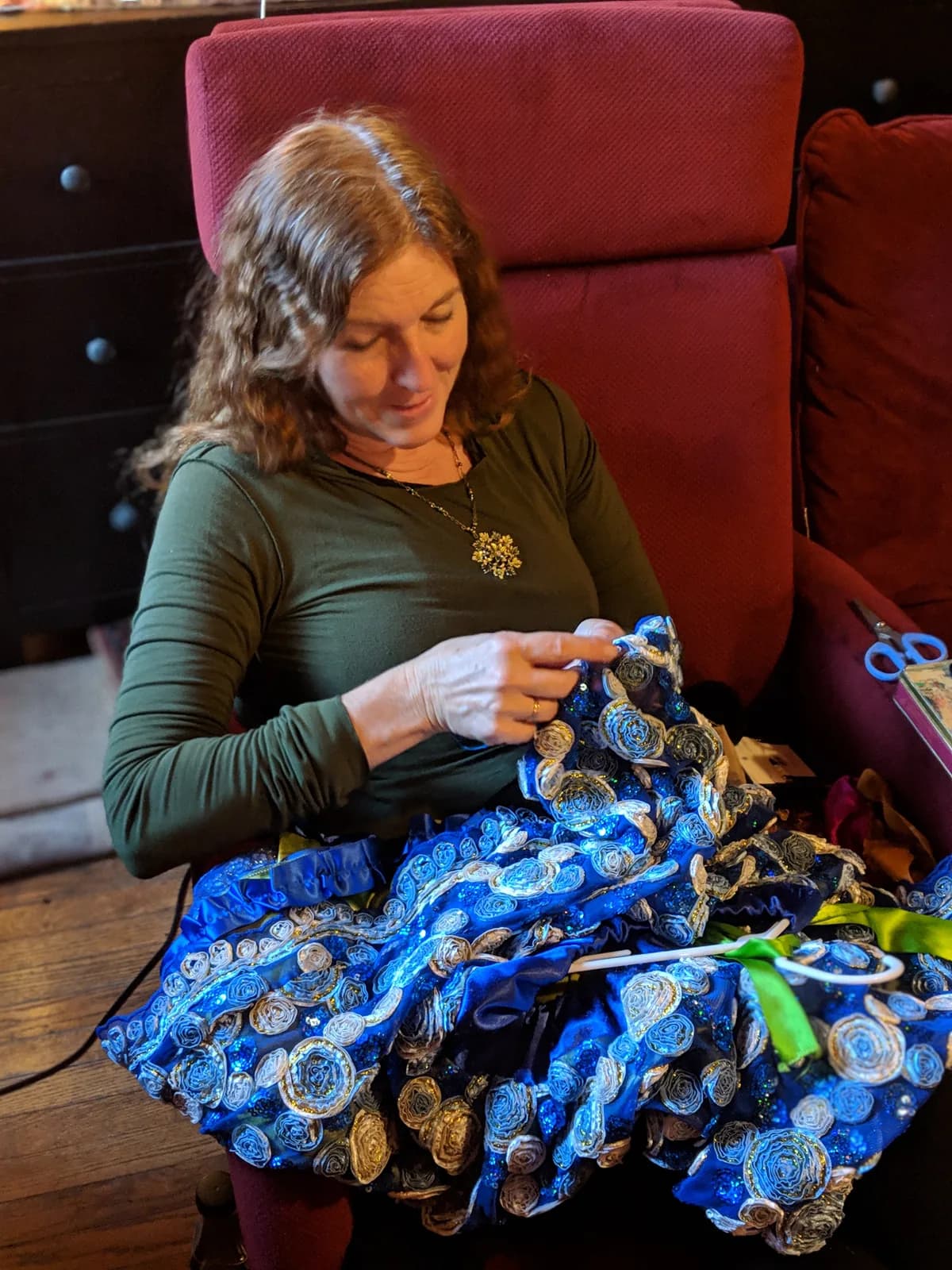Kim Snyder, lead costume designer, sits in a red armchair hand-sewing a blue and gold costume.