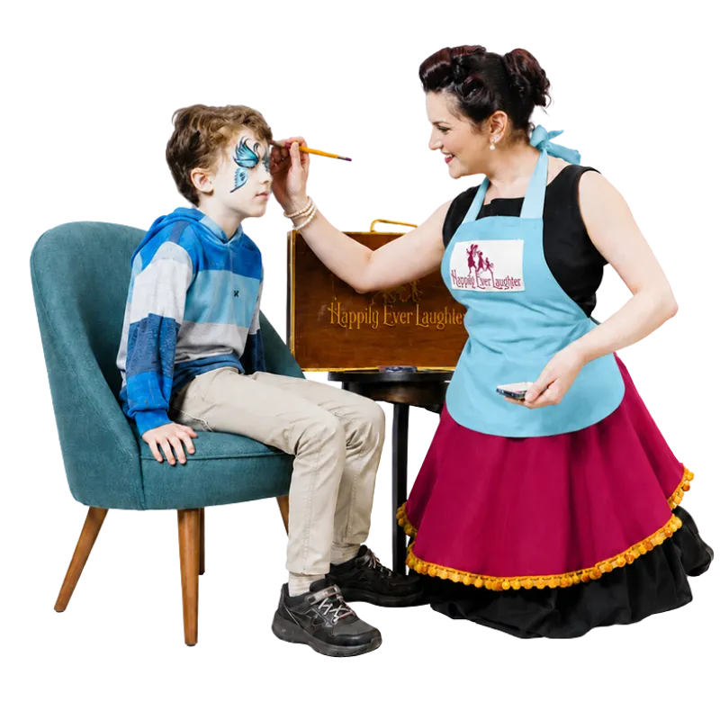 A Happily Ever Laughter face painter in a branded apron painting a blue butterfly design on a smiling child seated in an armchair.