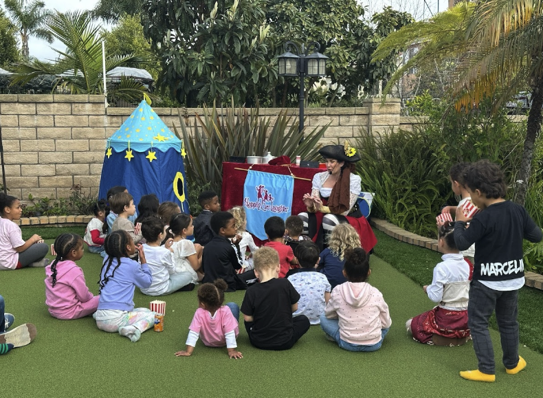 Children seated on artificial turf watching a pirate-themed puppet show outdoors.