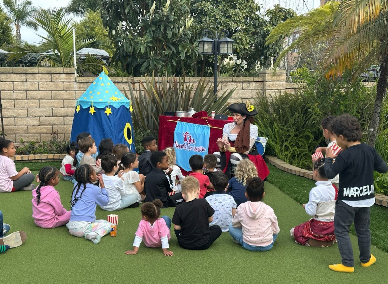 Children seated on artificial turf watching a pirate-themed puppet show outdoors.