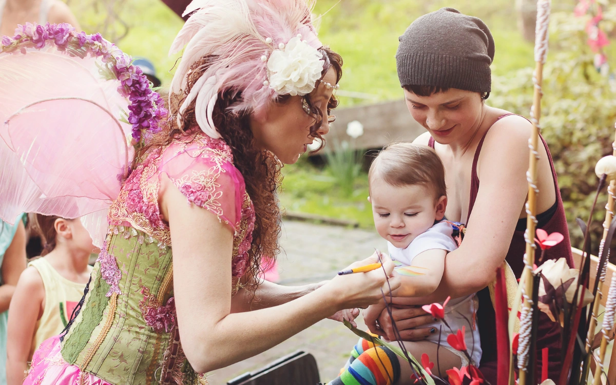 A Happily Ever Laughter face painter in a fairy costume painting a baby's arm while a parent holds them, with children watching.