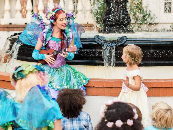 Happily Ever Laughter fairy performer playing the ukulele and singing to a group of children near a fountain, creating an enchanting Easter celebration at a country club.