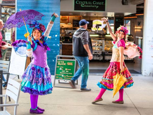 Happily Ever Laughter entertainers bringing magic and joy to a marketplace, dressed in vibrant costumes with umbrellas and wings, ideal for creating memorable experiences at public events or parties.
