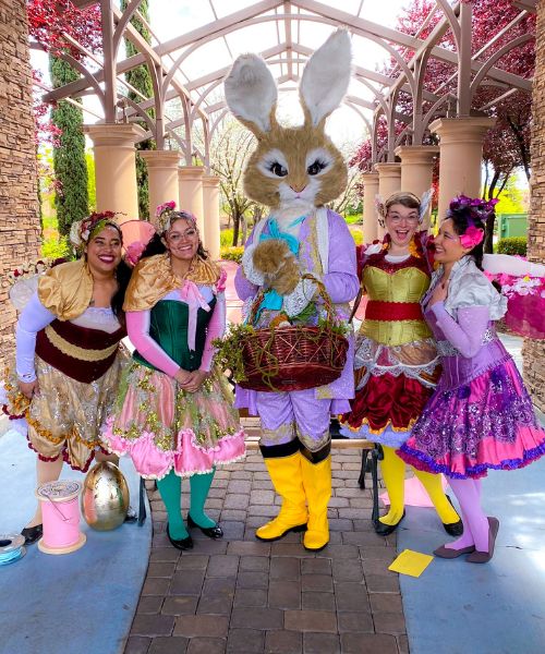 Group of performers from Happily Ever Laughter dressed in whimsical fairy costumes, posing with a life-sized Easter Bunny in a vibrant outdoor setting with spring-themed decorations. Perfect for Easter entertainment, kids' parties, and holiday events in California.